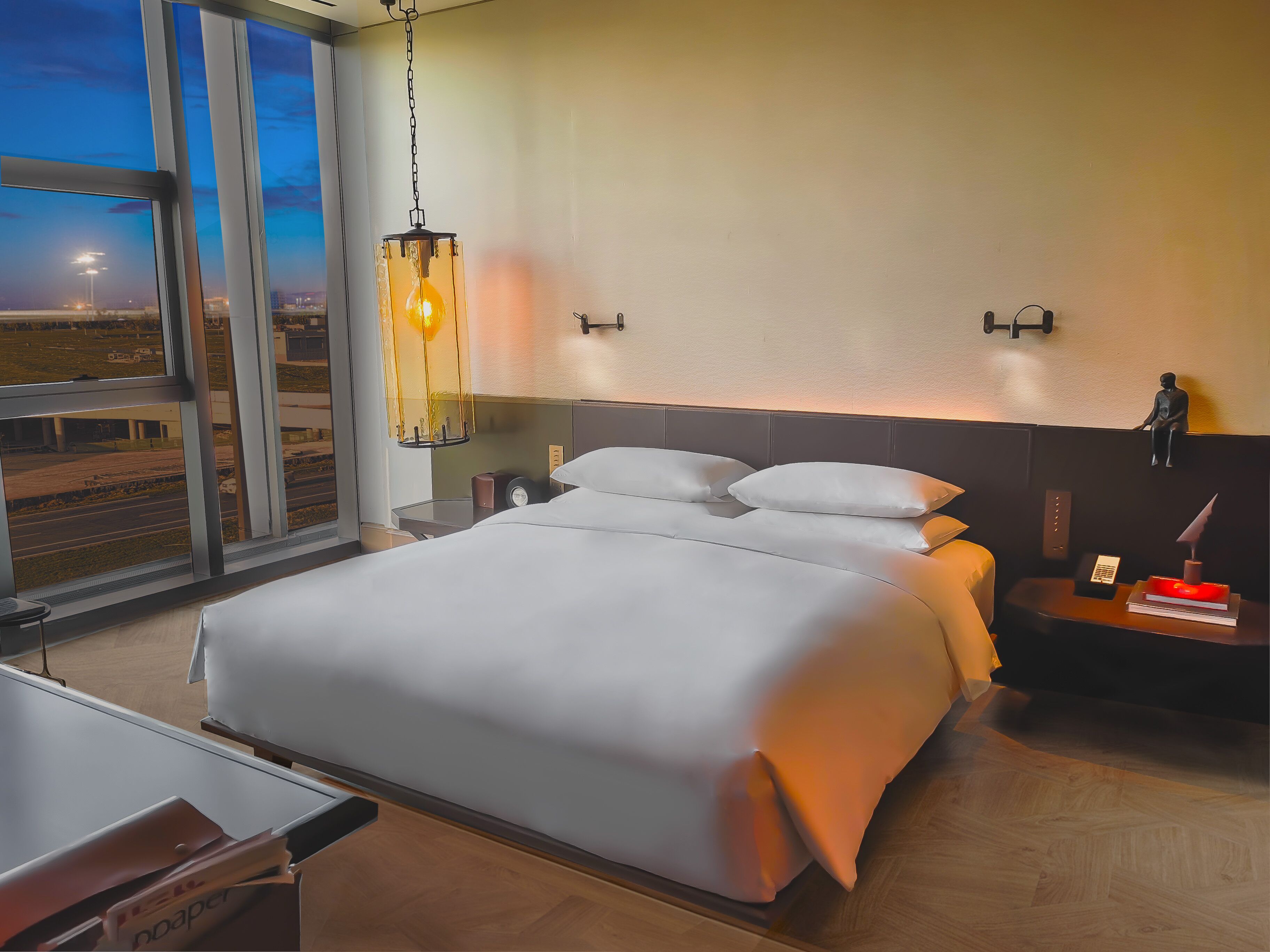 Una habitación de hotel moderna con una cama grande, mesitas de noche, un escritorio y vistas al aeropuerto.
