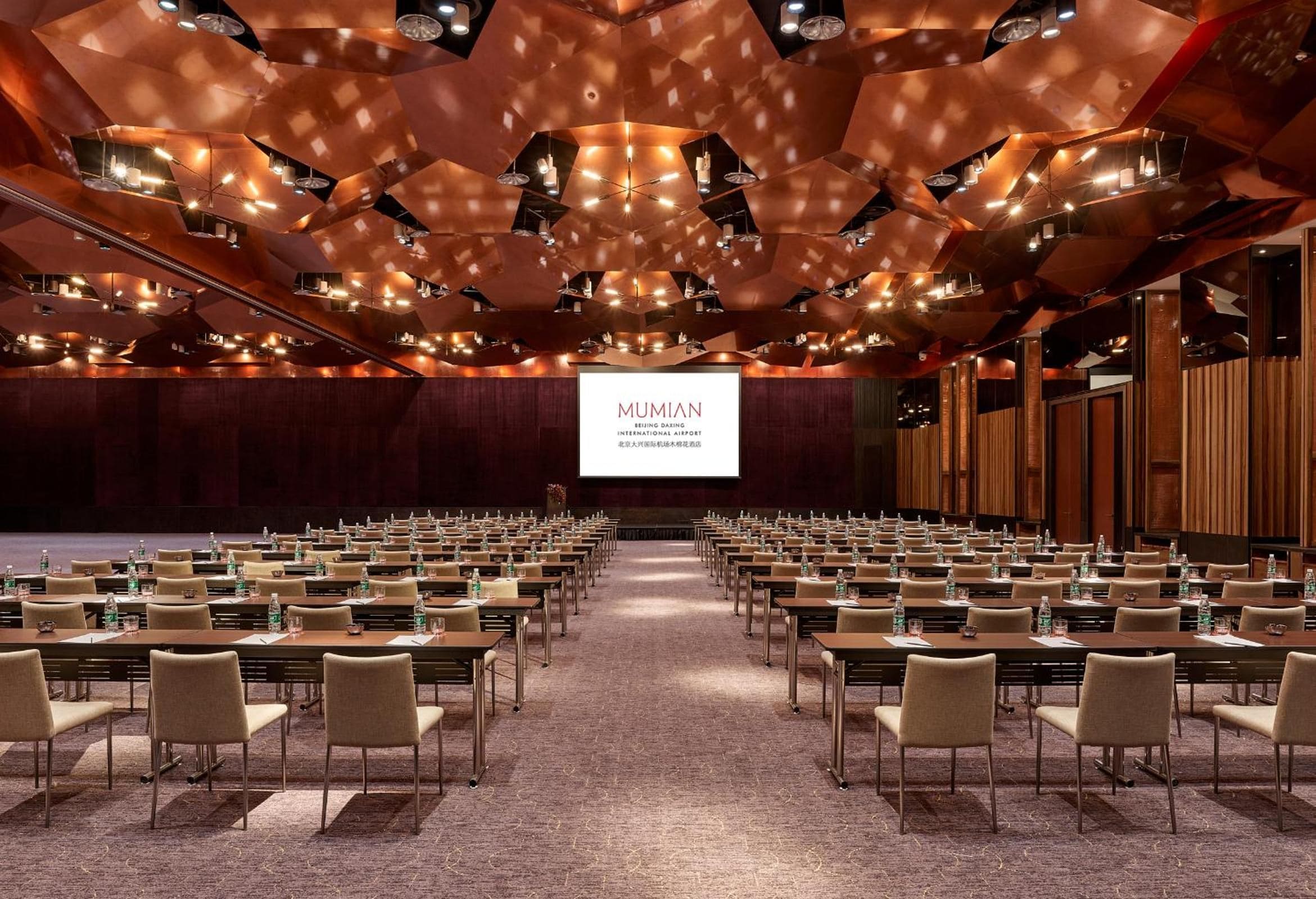 A conference room with rows of tables and chairs, a large screen at the front, and a ceiling with geometric patterns.