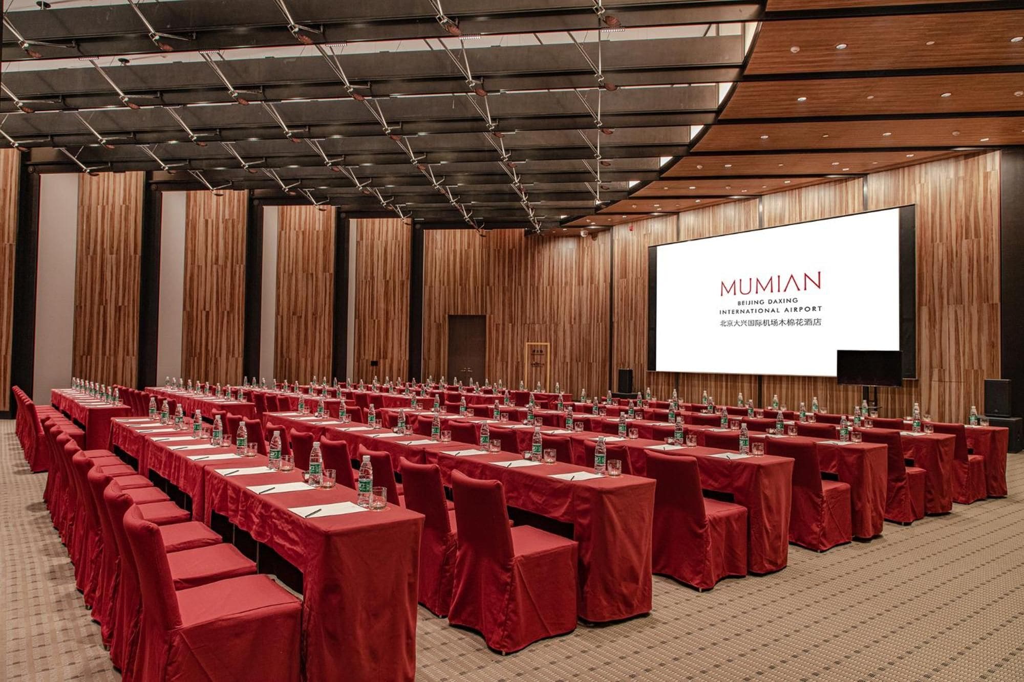 A conference room with rows of tables, red chairs, and a large screen displaying 'MUMIAN' text.