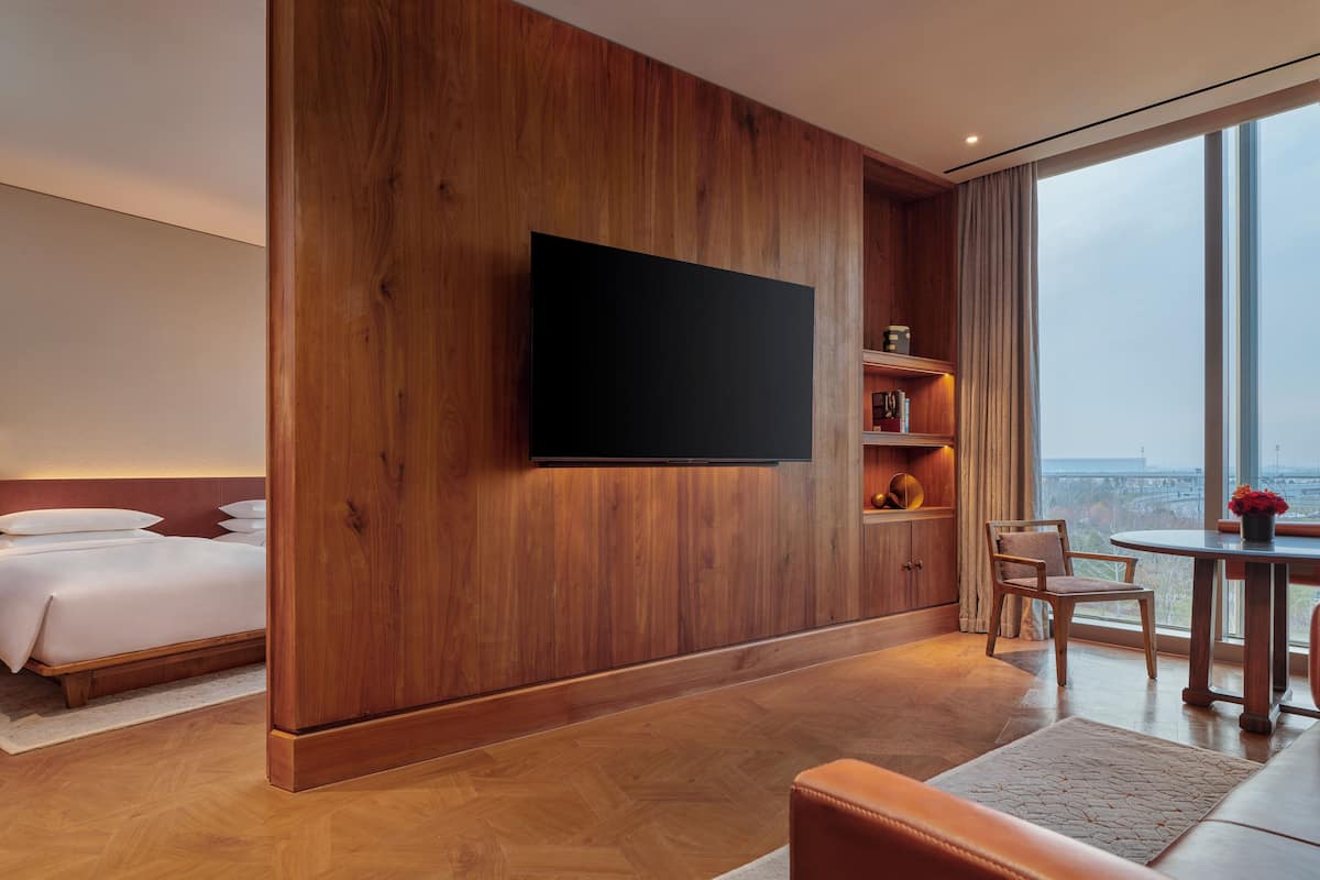 Ein modernes Hotelzimmer mit großem Fernseher an einer Holzwand, einem Bett, einer Sitzecke mit Tisch und Blick auf die Stadt durch große Fenster.