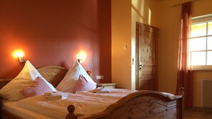 Una cama bien hecha con almohadas y un cabecero de madera, una lámpara fijada a la pared y una puerta con un ventanal a la derecha.