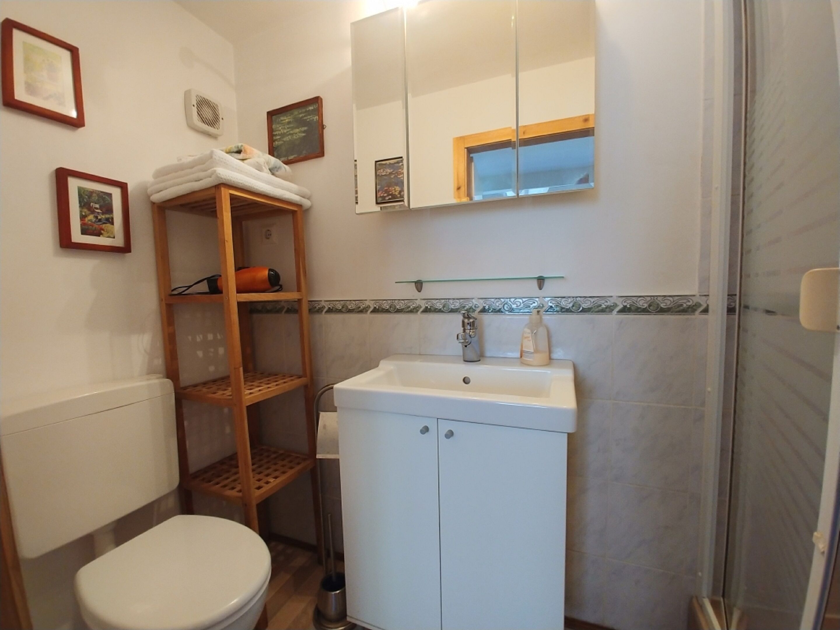 A bathroom with a toilet, a white sink with a mirror above it, a shelf with towels, and a picture frame on the wall.