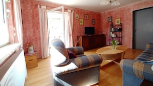 Una sala de estar con un sofá, una mesa de centro y una televisión. Hay una estantería y una puerta en la habitación.