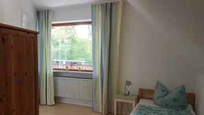 Une chambre avec une armoire en bois, un lit recouvert d’une couette à motifs, une table de chevet avec une lampe et une fenêtre avec des rideaux.