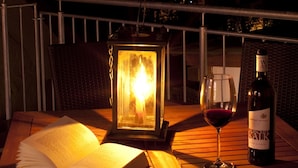 Una mesa de madera con un libro abierto, una copa de vino tinto, una botella de vino y un farol encendido en un balcón de noche.