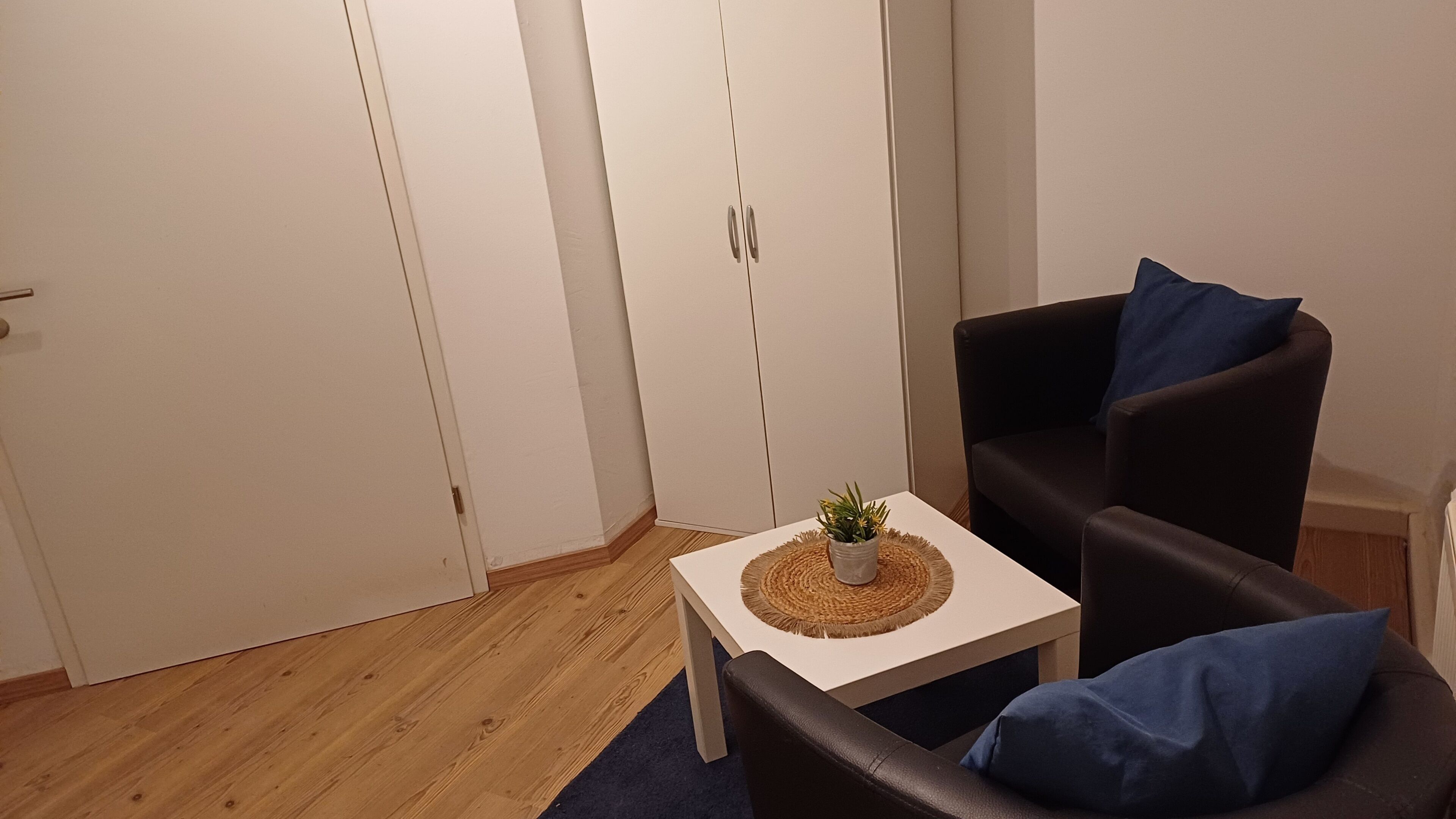 A small room with a wooden floor, a white table with a plant, a black armchair with a blue pillow, and a white wardrobe with a door.