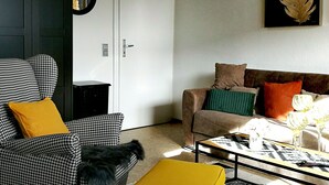 A living room with a houndstooth armchair, a sofa with colorful cushions, a coffee table with decorative items, and a door with a mirror. - Ferienwohnungen Stadler (Roedental)