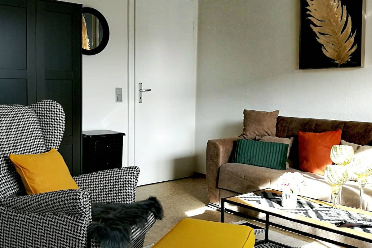 Een woonkamer met een ruitjes fauteuil, een bank met kleurrijke kussens, een salontafel met decoratieve objecten, en een deur met een spiegel.