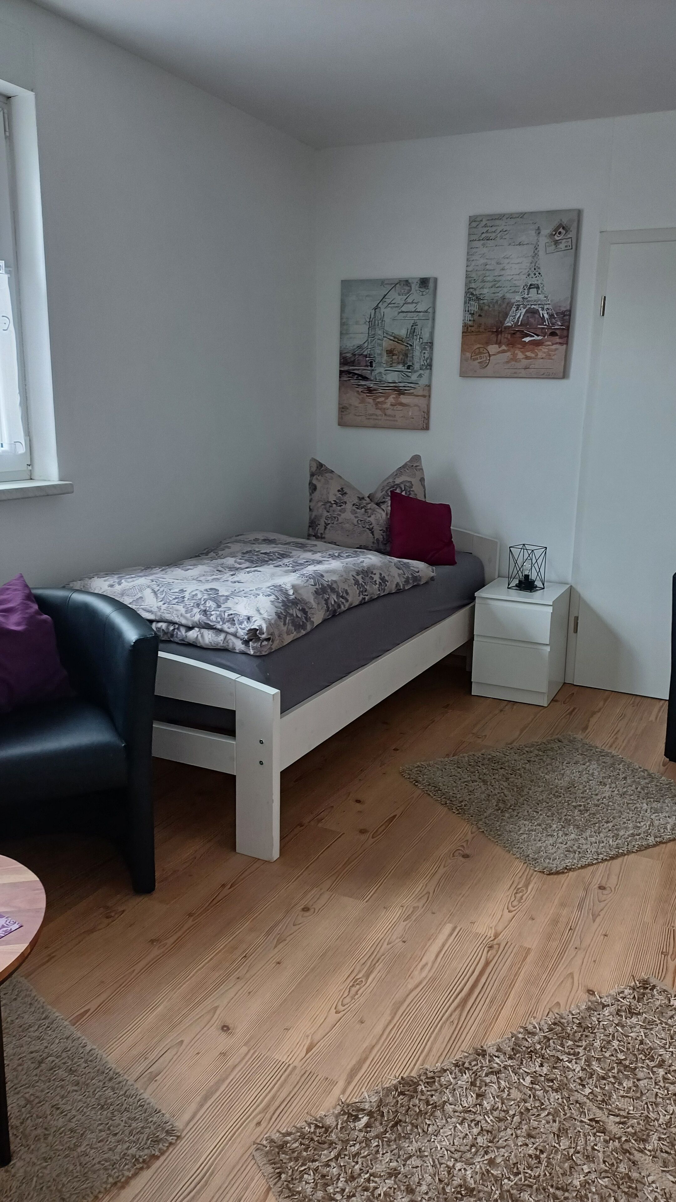 A small, neatly arranged bedroom with a single bed, a bedside table, a nightstand, and a window.