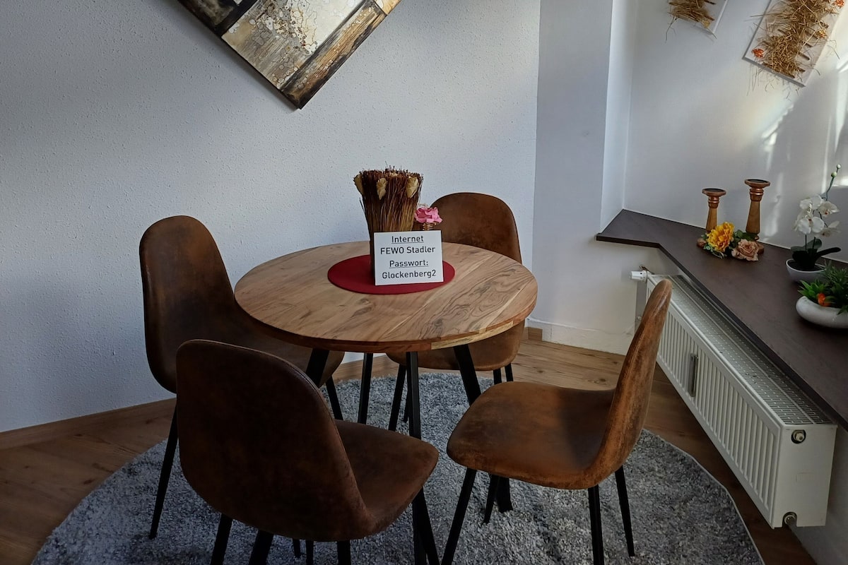 Een eetgedeelte met een ronde houten tafel, vier bruine stoelen, een bord met een internetsleutel en een raam met een teddybeer op de vensterbank.