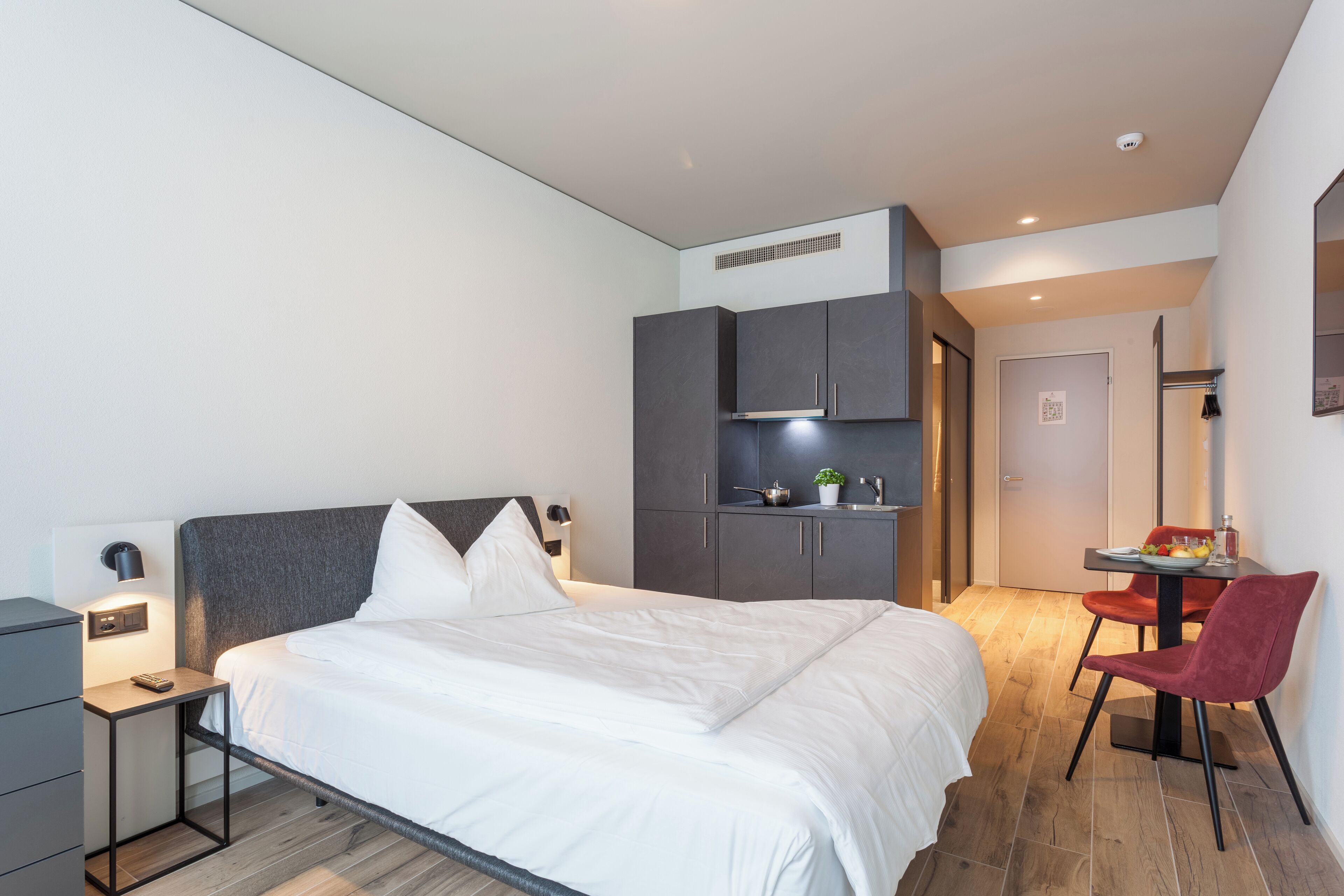 Een moderne hotelkamer met een groot bed, een bureau en een stoel. Er is een kleine tafel met een plant en een schaal fruit. De kamer heeft een grijze inloopkast en een witte deur.