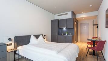 A modern hotel room with a large bed, a desk, and a chair. There is a small table with a plant and a fruit bowl. The room has a gray wardrobe and a white door.