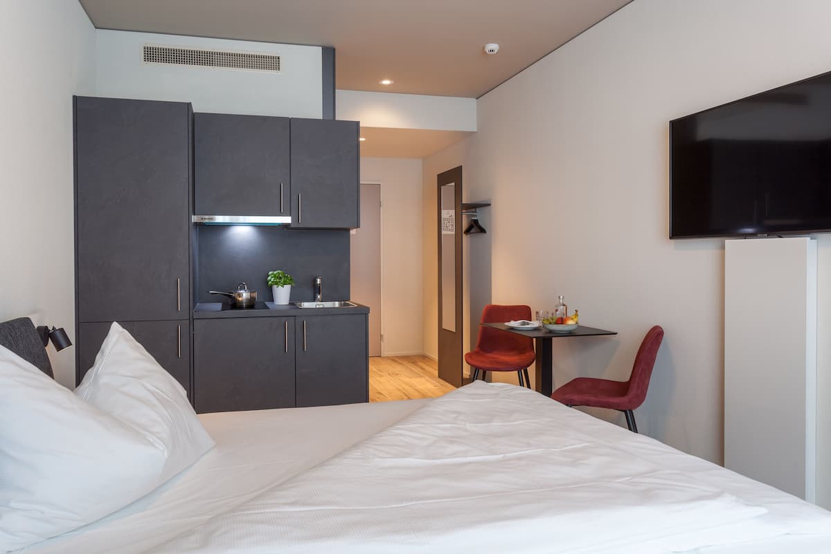 A modern hotel room with a bed, a small kitchenette, a TV, and a red chair.