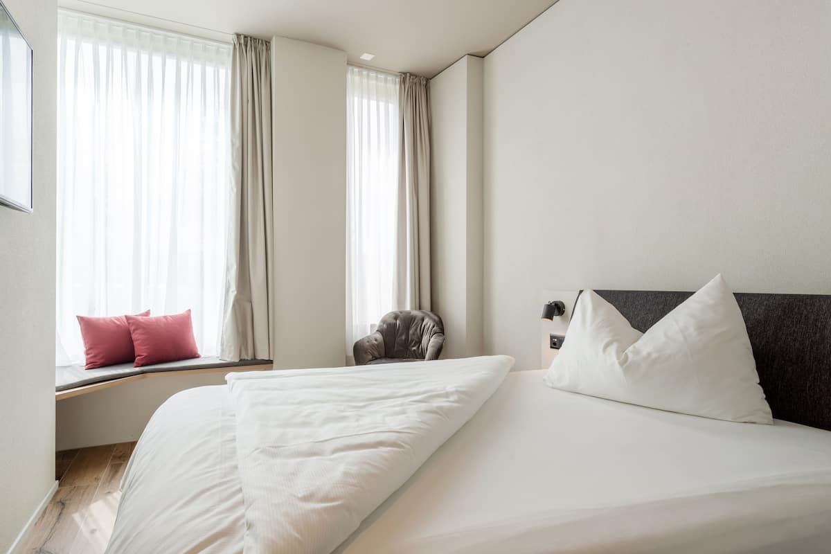 A hotel room with a large bed, a window seat with a chair, and a view of the outside.