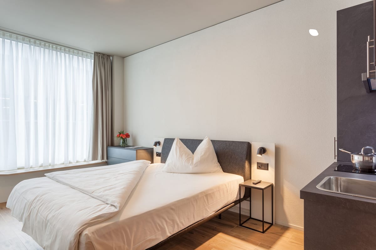 A modern hotel room with a large bed, bedside table, and a built-in kitchen area with a sink and stove.