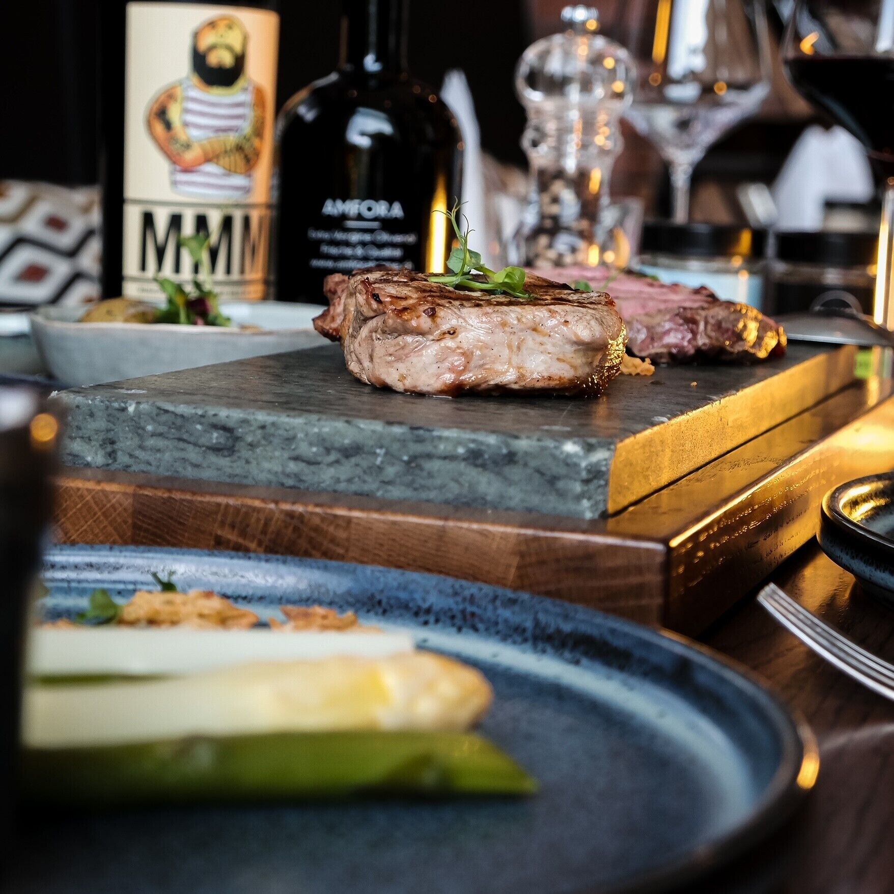 A steak on a wooden board, a bottle of Amfora wine, and a glass of red wine on a table.