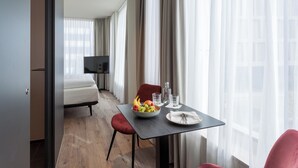 Habitación de hotel con una mesa de comedor dispuesta con fruta, vasos y un plato. Se ve una cama y un televisor al fondo.