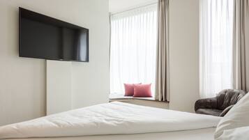 A modern hotel room with a large bed, a flat-screen TV mounted on the wall, a window with sheer curtains, and a window seat with a pink pillow.