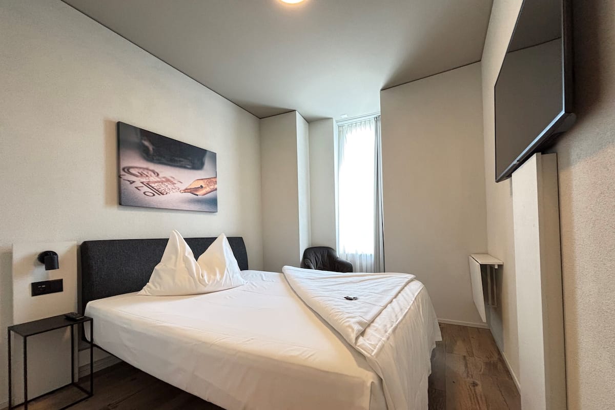 A hotel room with a bed, a bedside table, a chair, a TV, and a window with curtains.