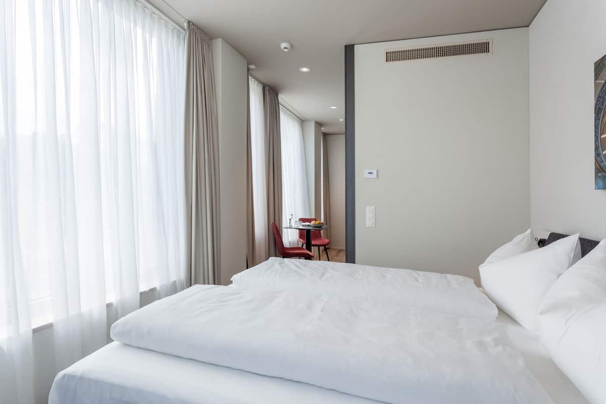 A hotel room with a large bed, a small table with a chair, and a window with curtains.