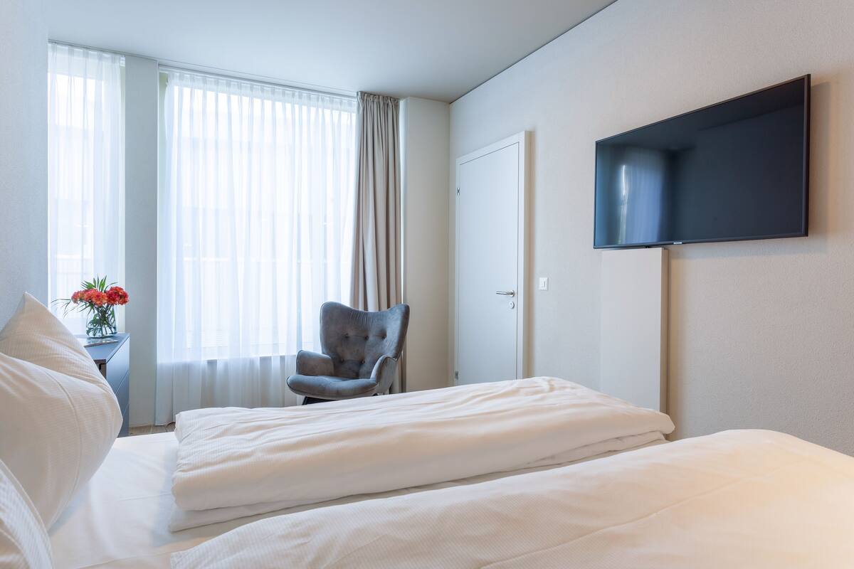 A hotel room with a bed, a chair, a TV mounted on the wall, and a window with curtains.