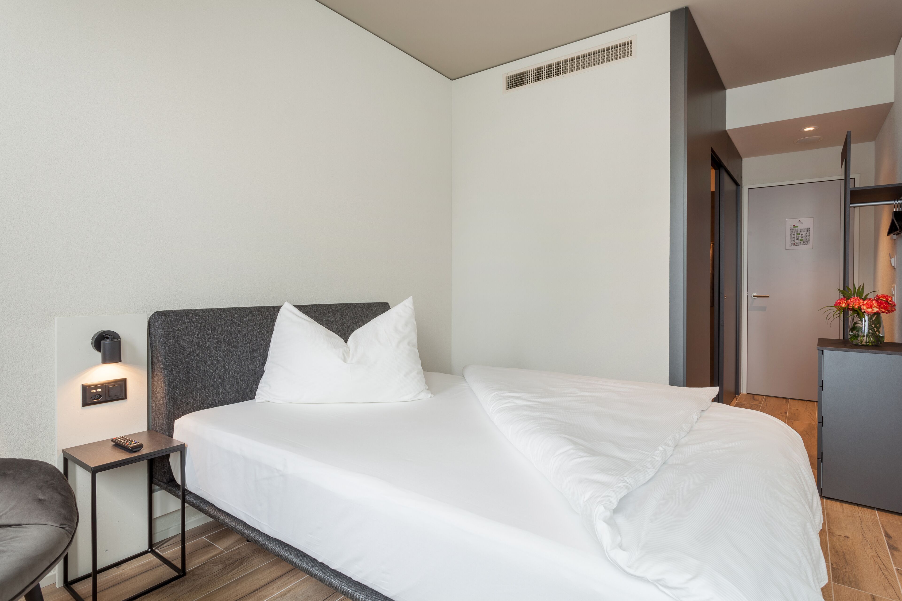 A modern hotel room with a neatly made bed, a bedside table with a lamp, and a small desk.