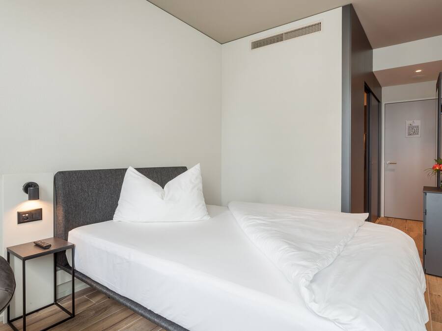 A modern hotel room with a neatly made bed, a bedside table with a lamp, and a small desk.