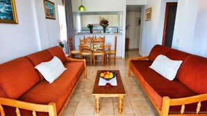 Una sala de estar con dos sofás rojos, una mesa de centro de madera con fruta, y una cocina al fondo.