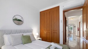 Un lit bien fait, avec du linge de lit blanc, un plaid vert et des serviettes pliées. On y trouve une armoire en bois surmontée d’un miroir, ainsi qu’une plante décorative fixée au mur.