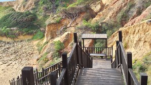 Un escalier en bois mène à une plateforme d’observation en bois, dotée d’une rambarde en métal, qui surplombe une côte rocheuse à la végétation clairsemée.