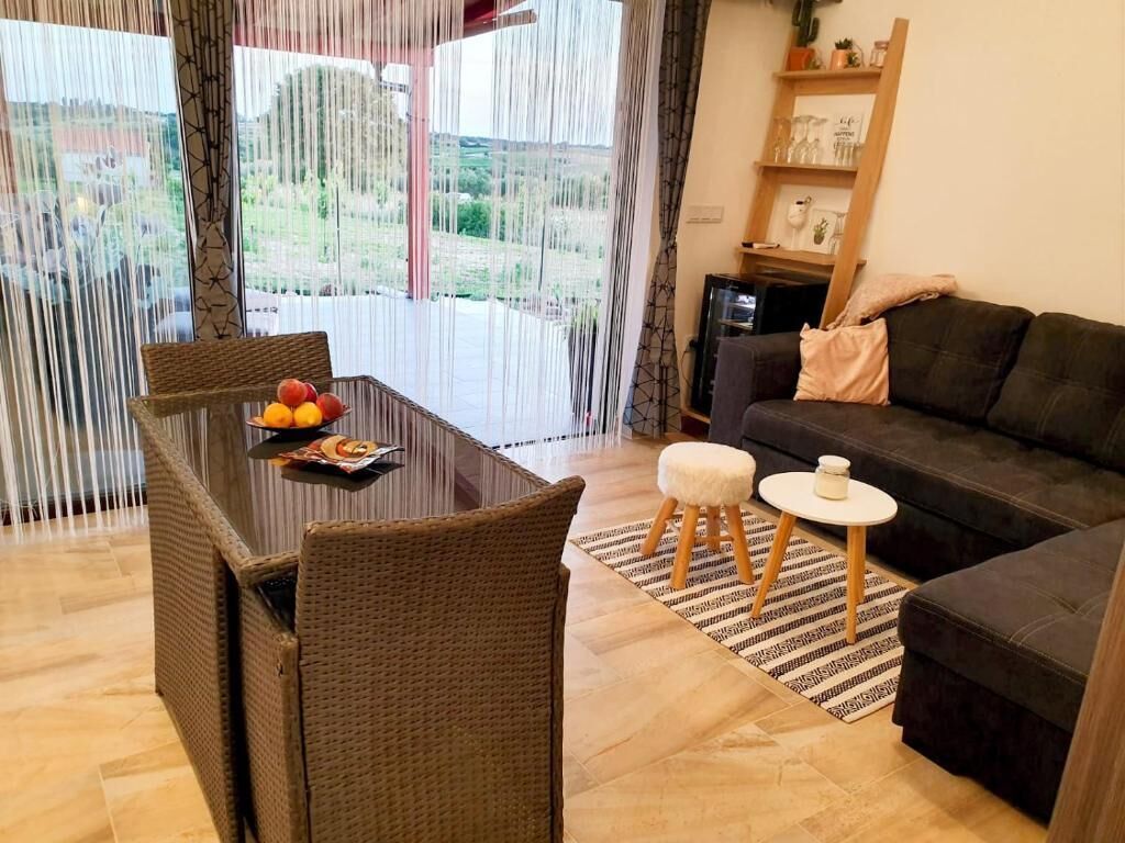 A living room with a dark sofa, a dining table with chairs, and a view through large windows.