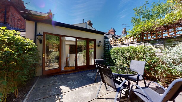 A patio area with a table and chairs, a sliding glass door leading to a garden, and a brick wall. - Princes Street | Oxford Holiday Let | 3 Beds & Sleeps 6 (Oxford)