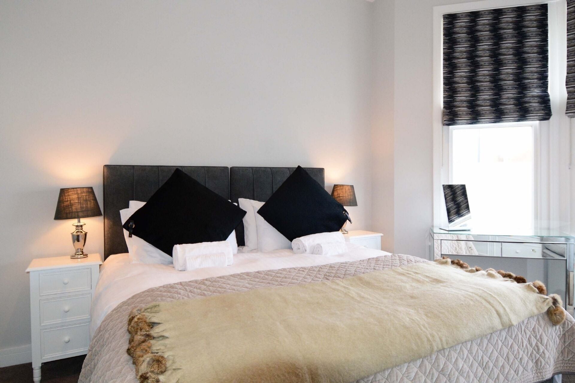 A neatly made bed with a quilted cover, two bedside tables with lamps, and a window with a patterned curtain.