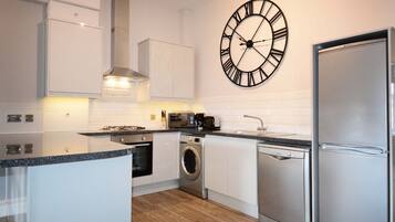 A modern kitchen with white cabinetry, a stainless steel refrigerator, a built-in oven, a washing machine, and a large wall clock.