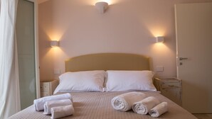 Un lit bien fait, avec du linge de lit blanc et des serviettes pliées, deux tables de chevet et des lampes fixées au mur.