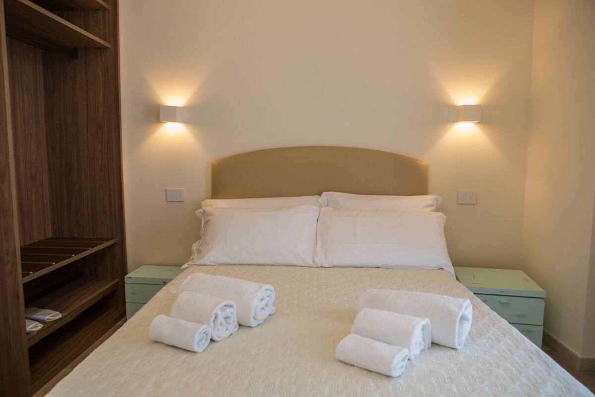 Una cama bien hecha con dos almohadas y toallas enrolladas sobre un edredón de color claro. A la izquierda hay un armario de madera y a la derecha una mesita de noche con una lámpara.