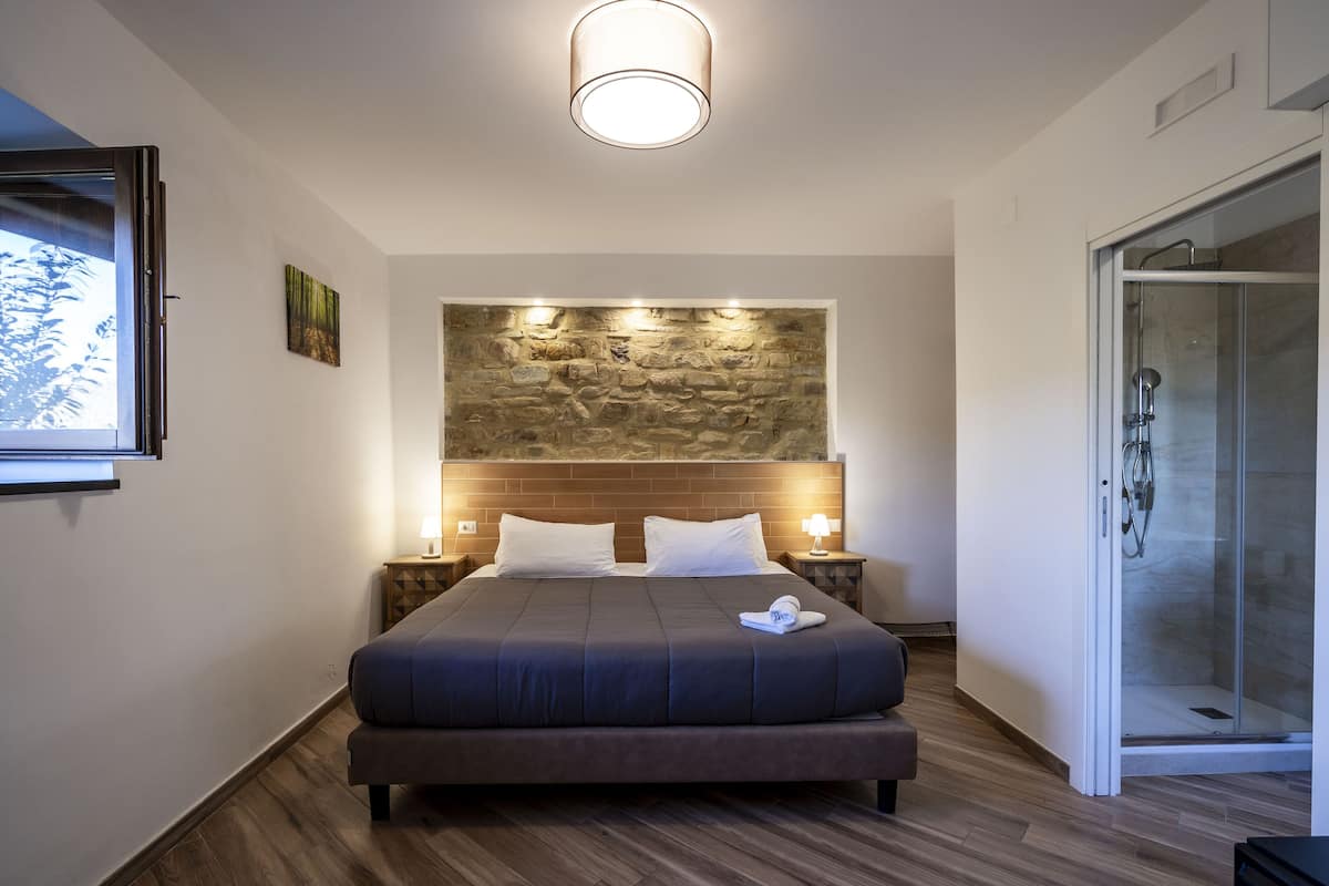 Ein modernes Hotelzimmer mit einem großen Bett, einer Wand aus Naturstein, einer Glasdusche und Blick ins Freie.