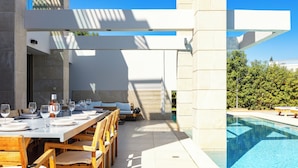 Un área de comedor al aire libre con piscina, sillas de madera y una mesa blanca dispuesta para una comida.