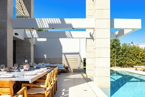 An outdoor dining area with a pool, wooden chairs, and a white table set for a meal.