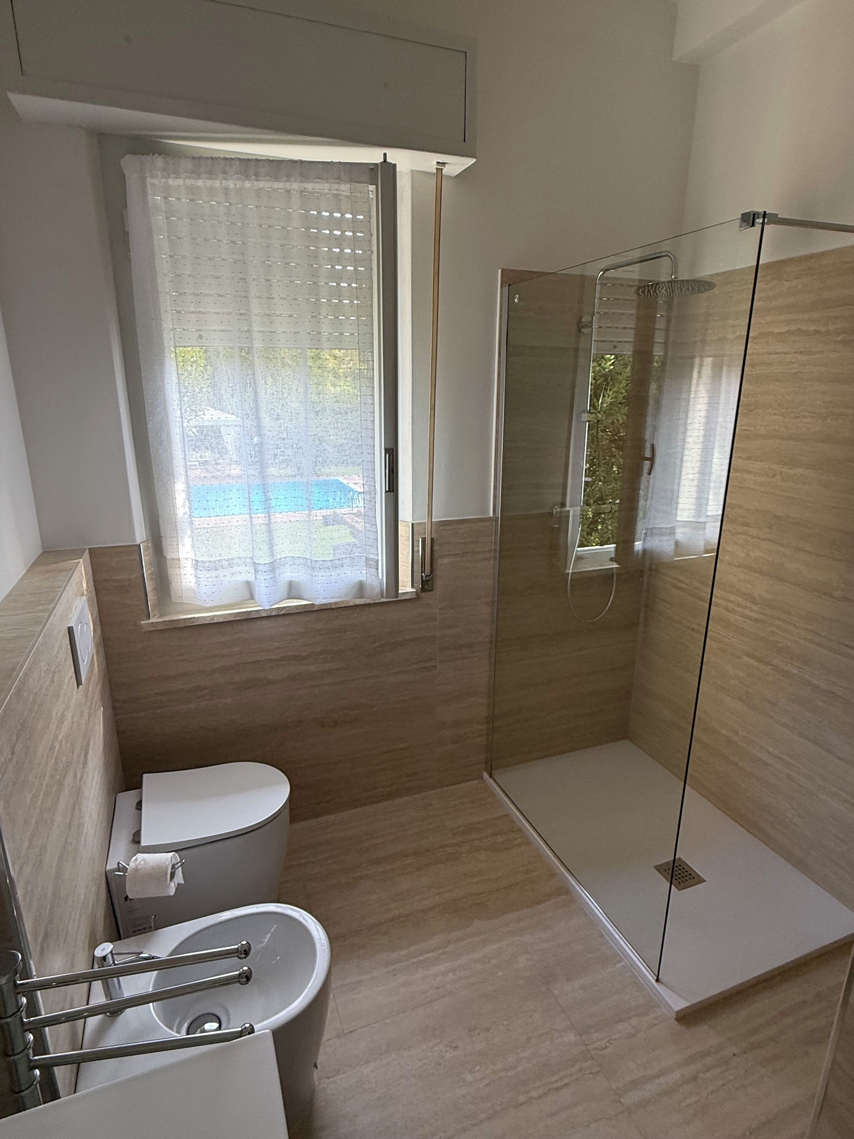 A modern bathroom with a toilet, a glass shower enclosure, and a window with a view of a pool.