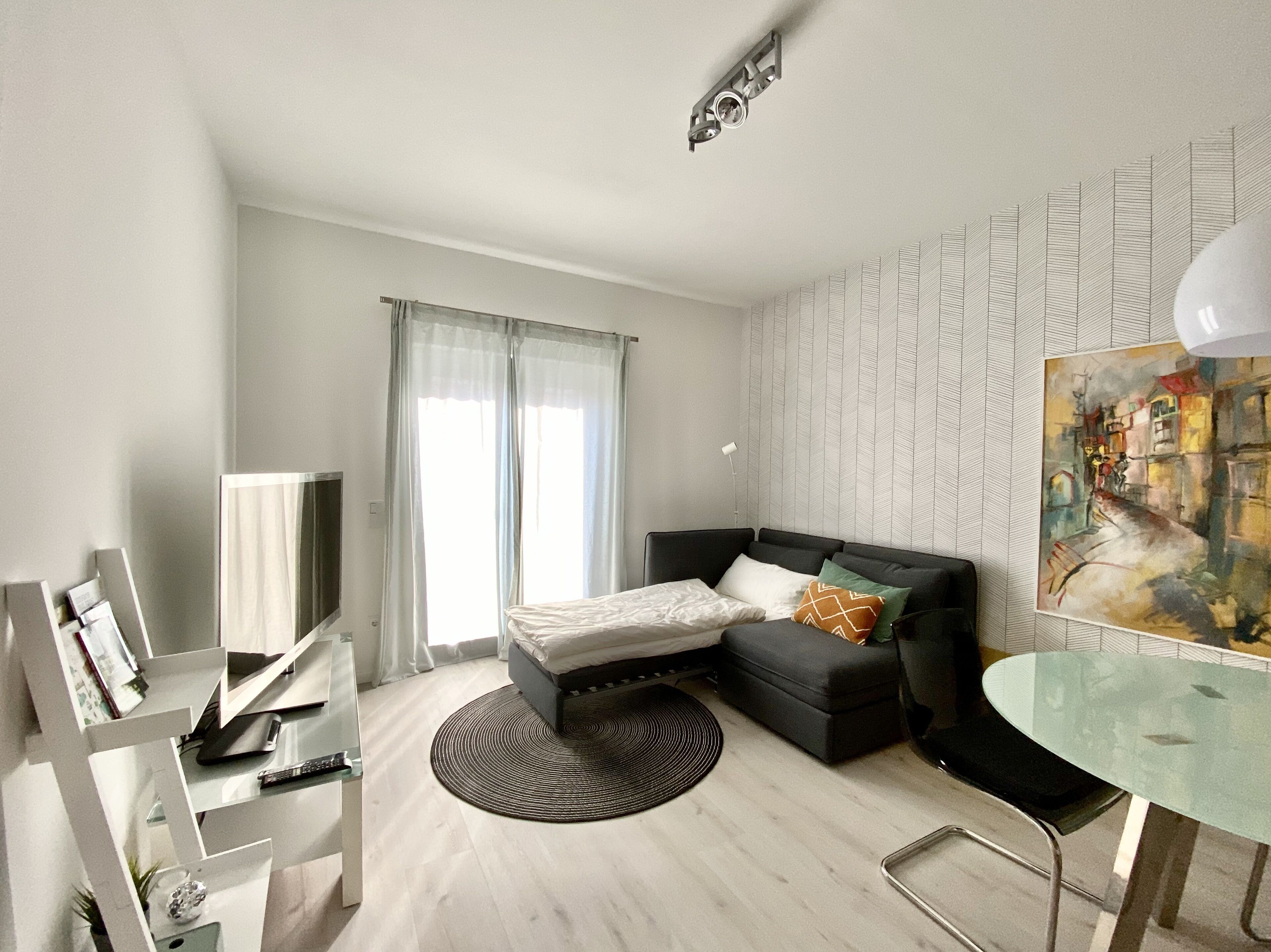 A modern, well-lit room with a bed, a sofa, a glass table, and a painting on the wall.