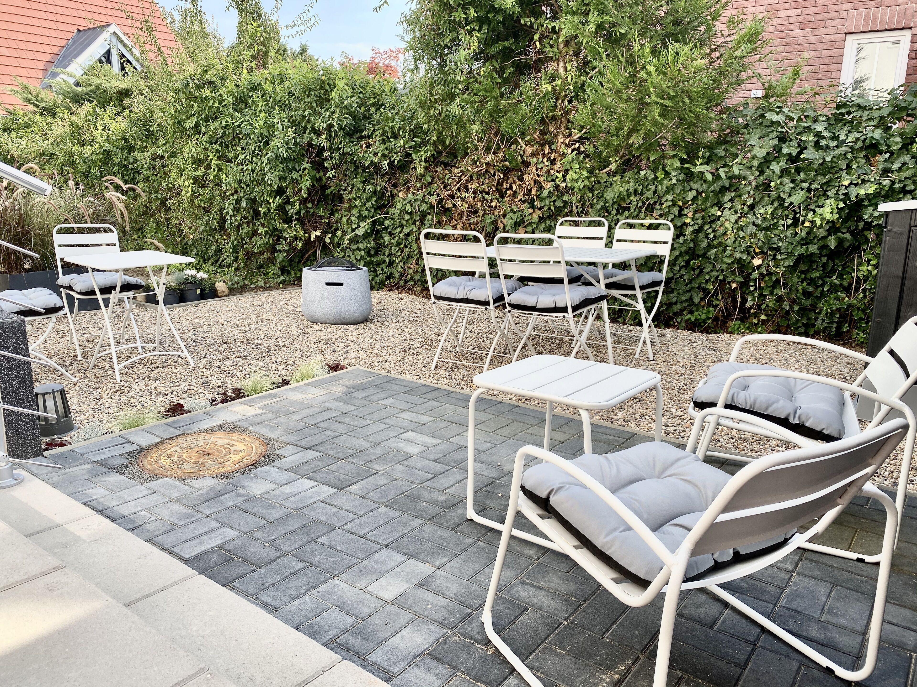 An outdoor patio area with white metal chairs and tables, surrounded by gravel and greenery.