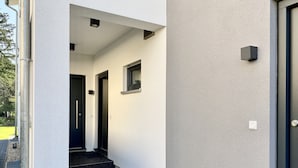Een smalle, buitenste gang met een zwarte deur en een raam, die leidt naar een donkere trap.