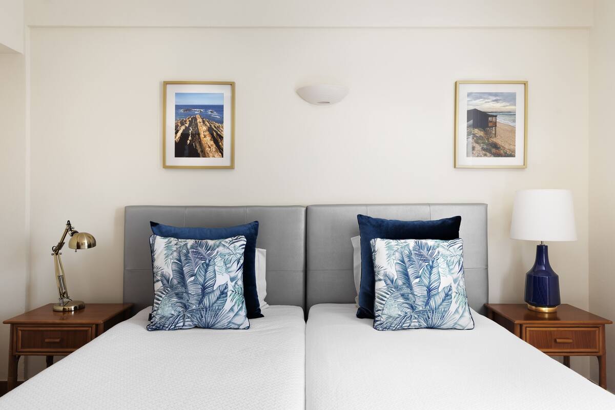 Un lit double avec des oreillers assortis, une table de chevet avec une lampe et deux tableaux encadrés au mur.