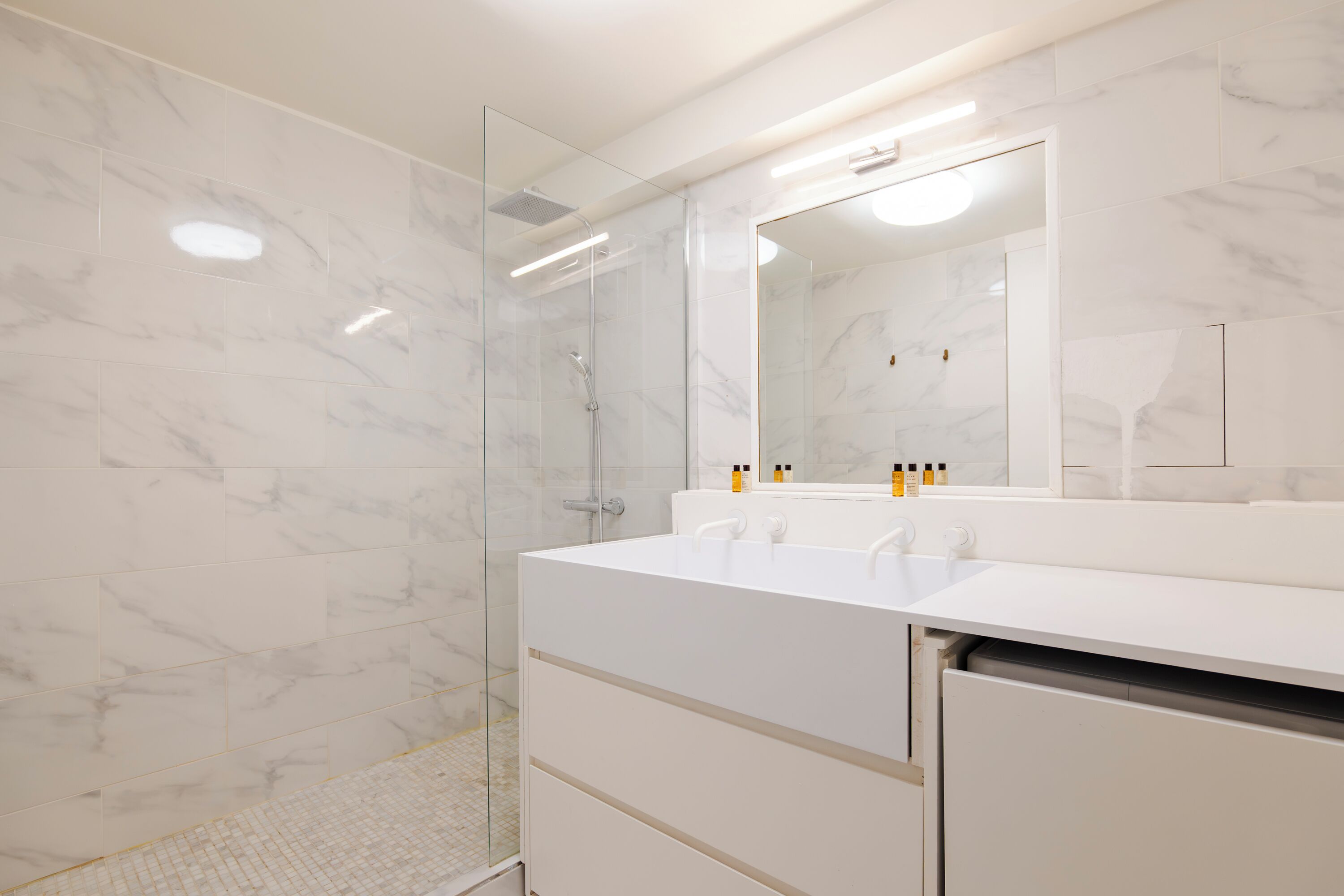 Une salle de bain moderne avec une douche à l’italienne en verre, des carreaux de marbre blanc et un grand miroir au-dessus du meuble-lavabo.