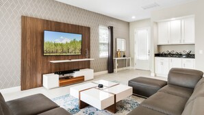 Een moderne woonkamer met een flatscreen televisie, een houten dressoir, een bank, een salontafel en een keuken met witte kastjes en een aanrecht.