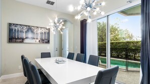 Een moderne eetkamer met een witte tafel en zwarte stoelen, een groot raam met uitzicht op een zwembad, en een plafondlamp.