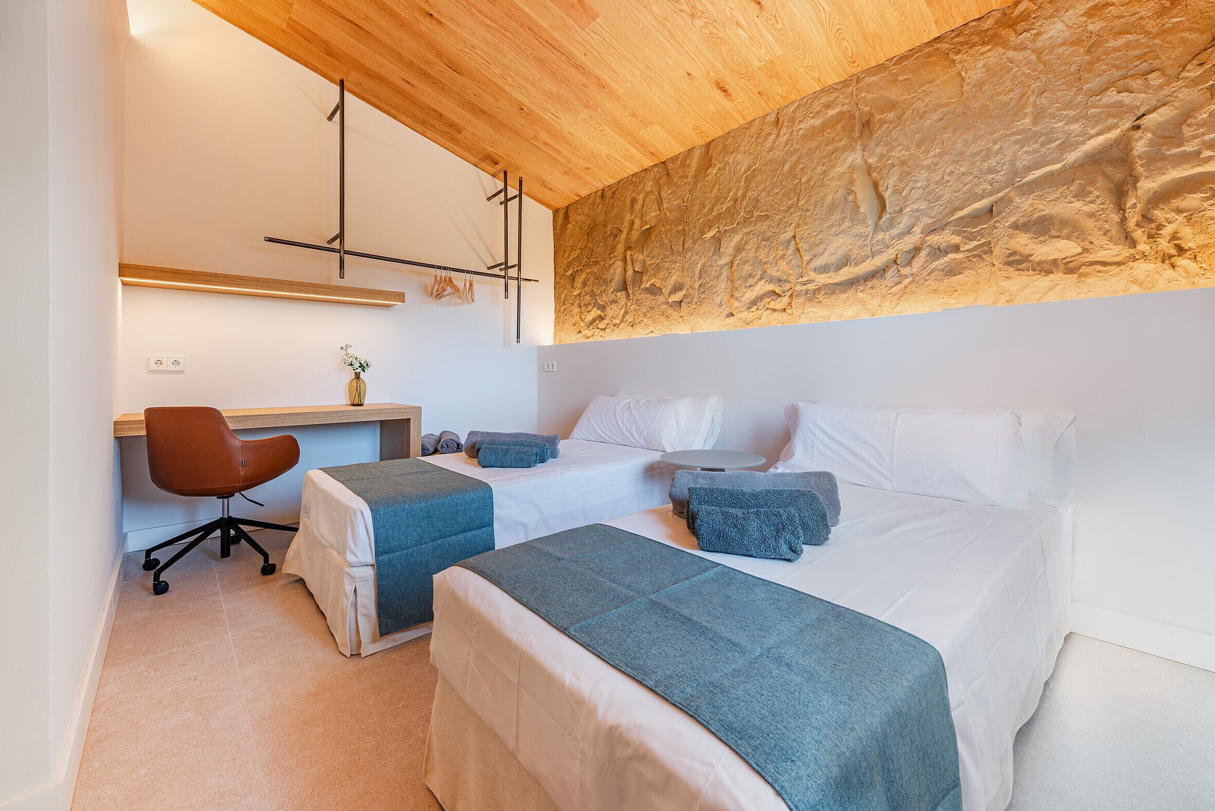 Habitación de hotel moderna con techo de madera, un escritorio con silla, dos camas con almohadas y una pequeña mesa redonda.