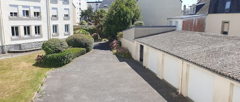 A narrow pathway flanked by buildings with balconies and a garden area with bushes and flowers.