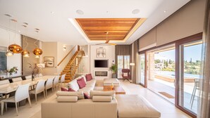 A modern living room with a large sofa, dining area, and a staircase leading to an upper level. - Villa Elea, New Deluxe Golf Villa in Aphrodite Hills - 6 Bedrooms, 7 Bathrooms.  (Kouklia)